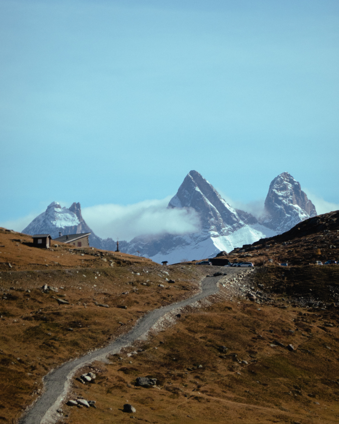 A winding mountain trail leads through brown alpine terrain towards snow-capped peaks under a bright blue sky.