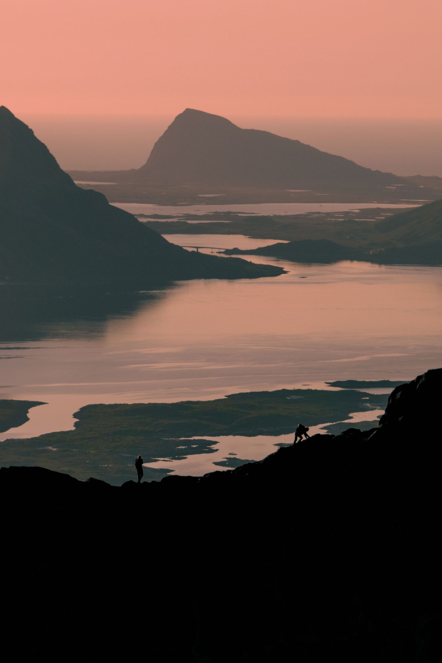 Silhouetted mountains and winding coastline at sunset create a dramatic landscape with pink and orange hues in the sky.