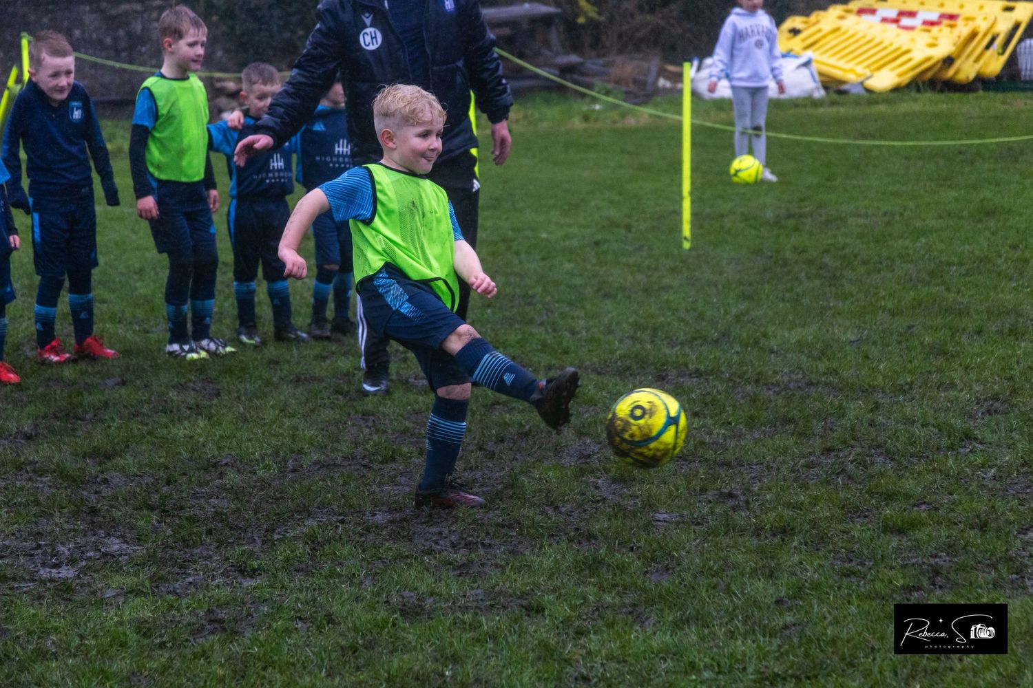 Children practice soccer drills with yellow cones and balls on muddy field.