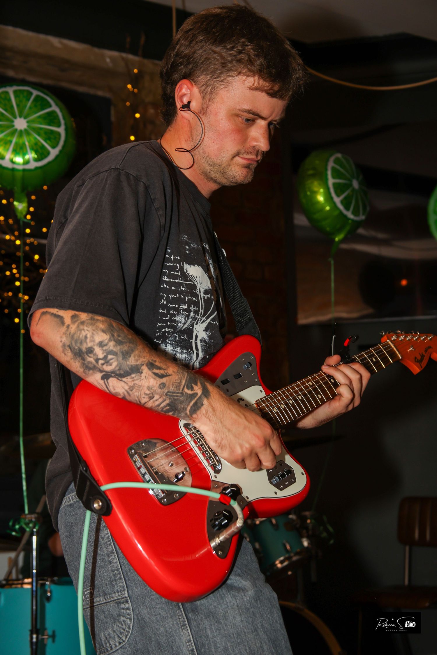 Guitarist playing red electric guitar under green neon lighting at small music venue.