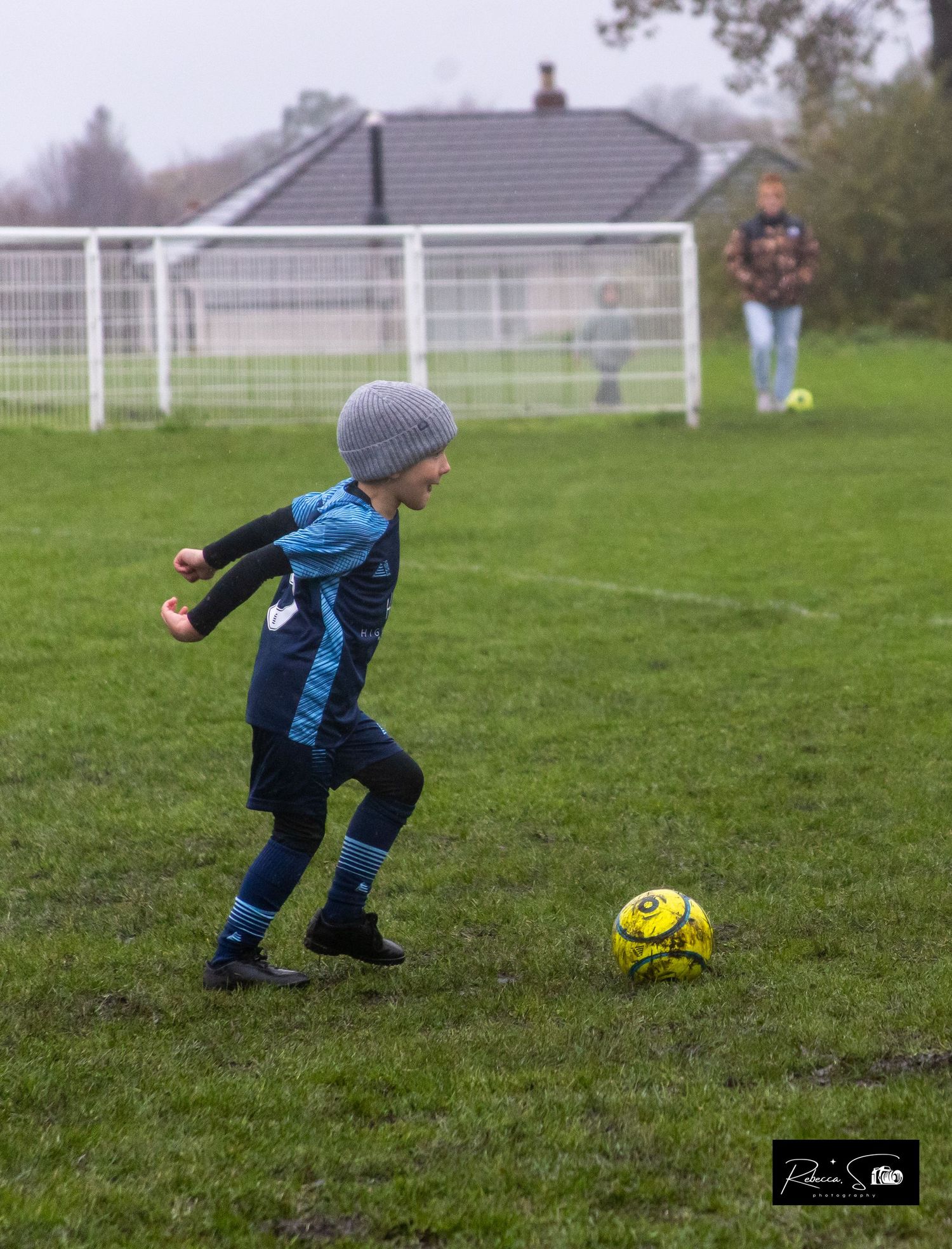 A young soccer player dribbles a yellow ball across a wet grass field.