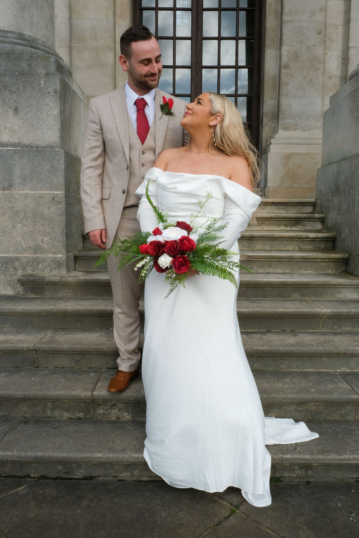 A couple shares a joyful moment on stone steps, with the bride wearing a white gown and holding a red bouquet.