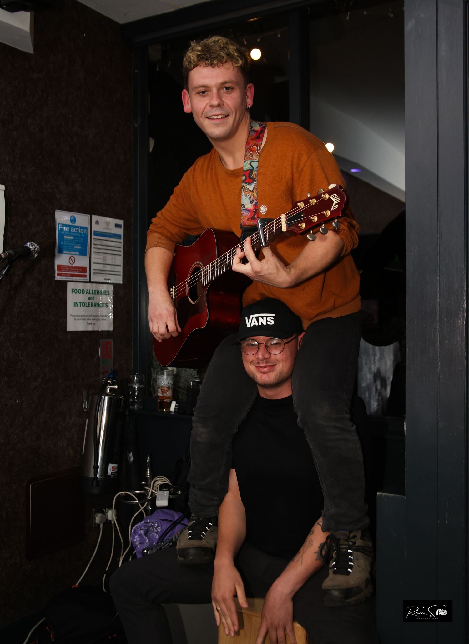 Two performers pose playfully on stage with acoustic guitar and sound equipment.