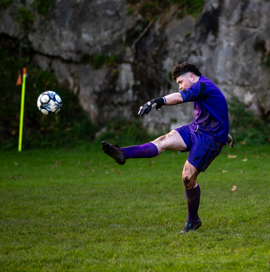 Soccer player in blue uniform performs acrobatic kick on grass field near rock wall.