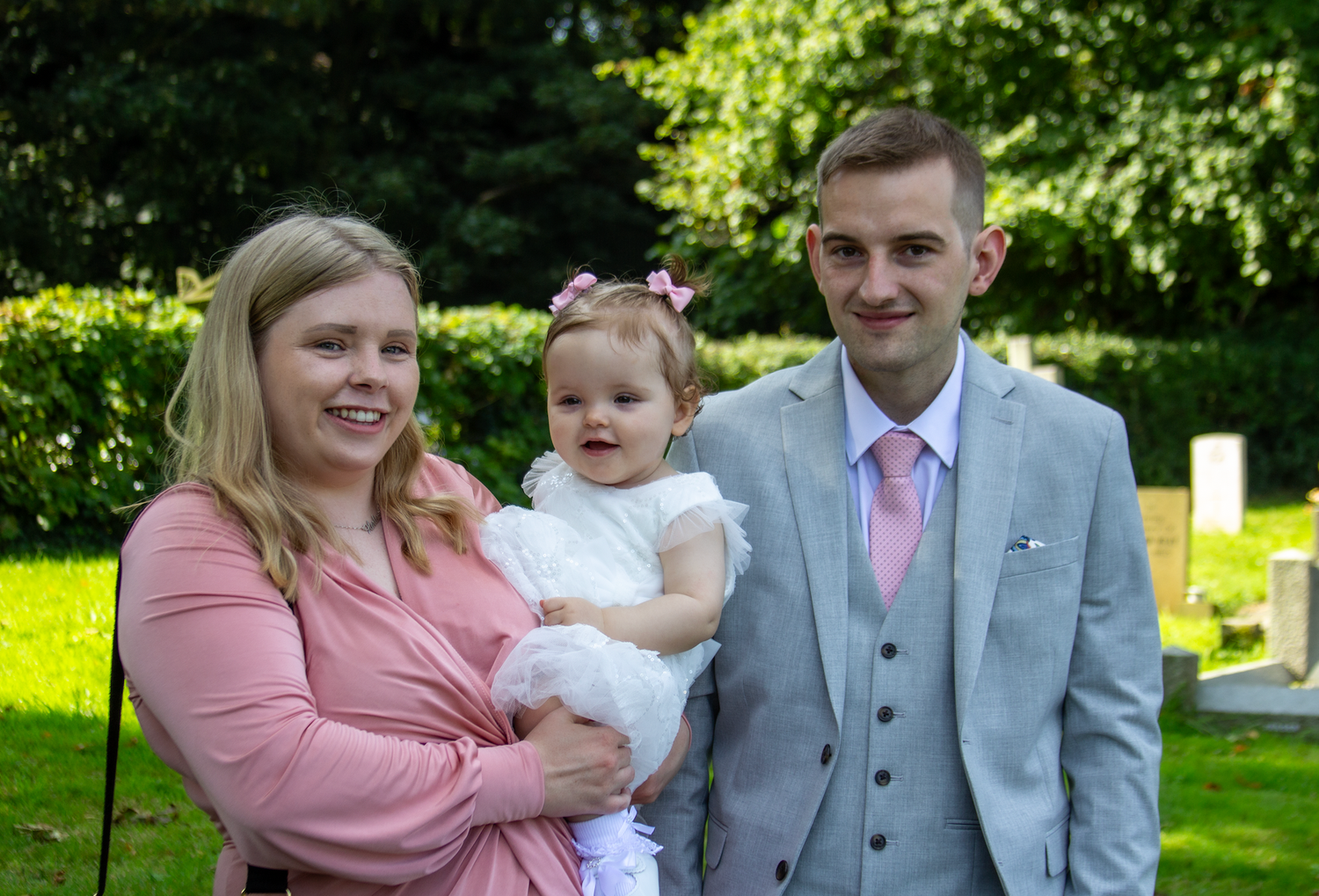 A wedding party poses together on a grassy lawn surrounded by trees and natural sunlight.