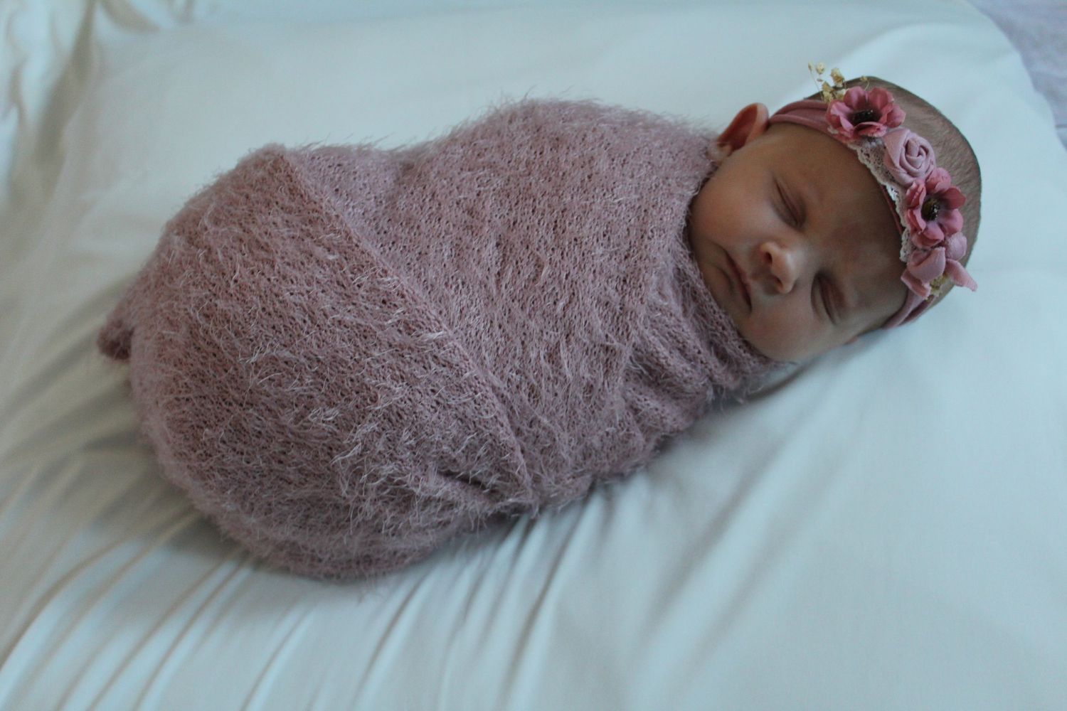Sleeping newborn baby swaddled in soft pink wrap on white bedding.