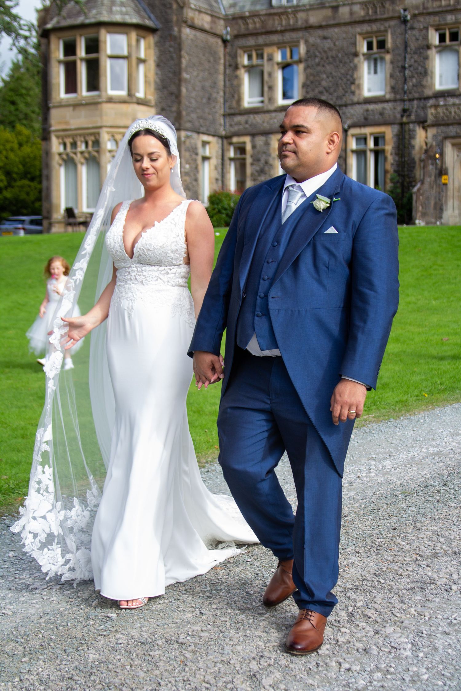 Wedding couple walking on a gravel path next to a stone building in elegant attire.