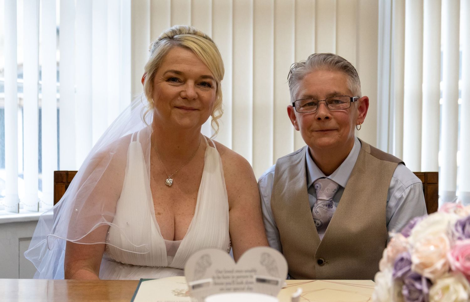 Wedding party signing official documents at a table with vertical blinds in background.