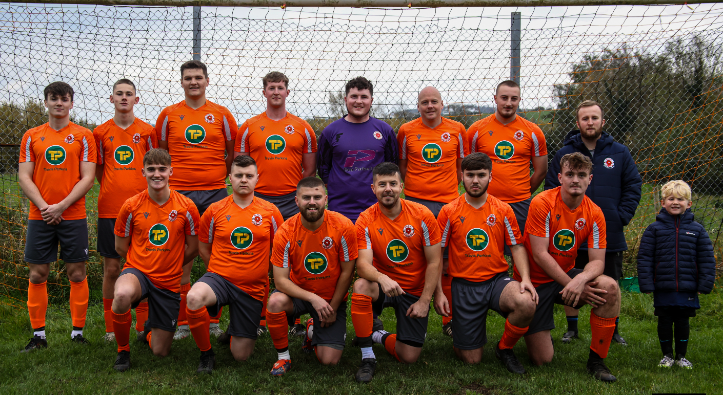 football team in orange jerseys poses together on grass field with goal net visible in background.