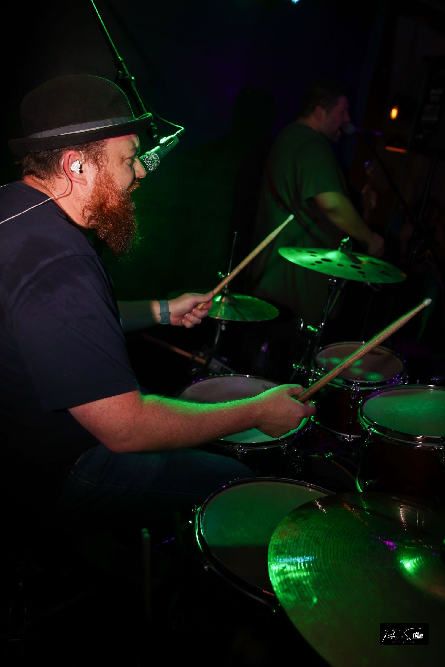 Drummer in black hat performing under green stage lighting.