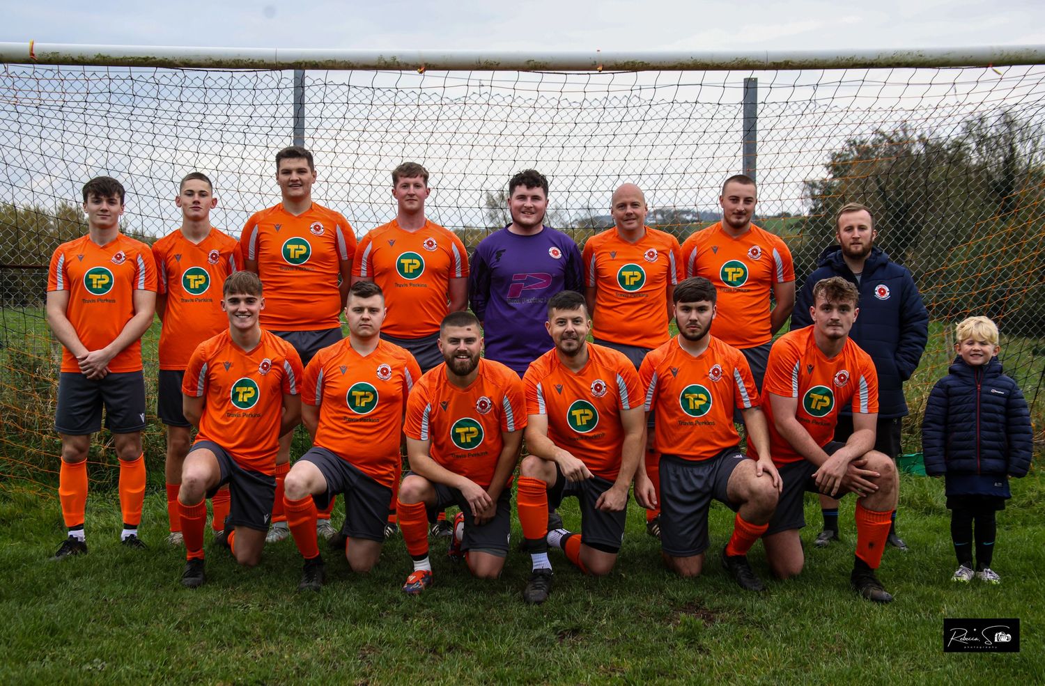 Soccer team in orange uniforms poses for group photo in front of goal net.