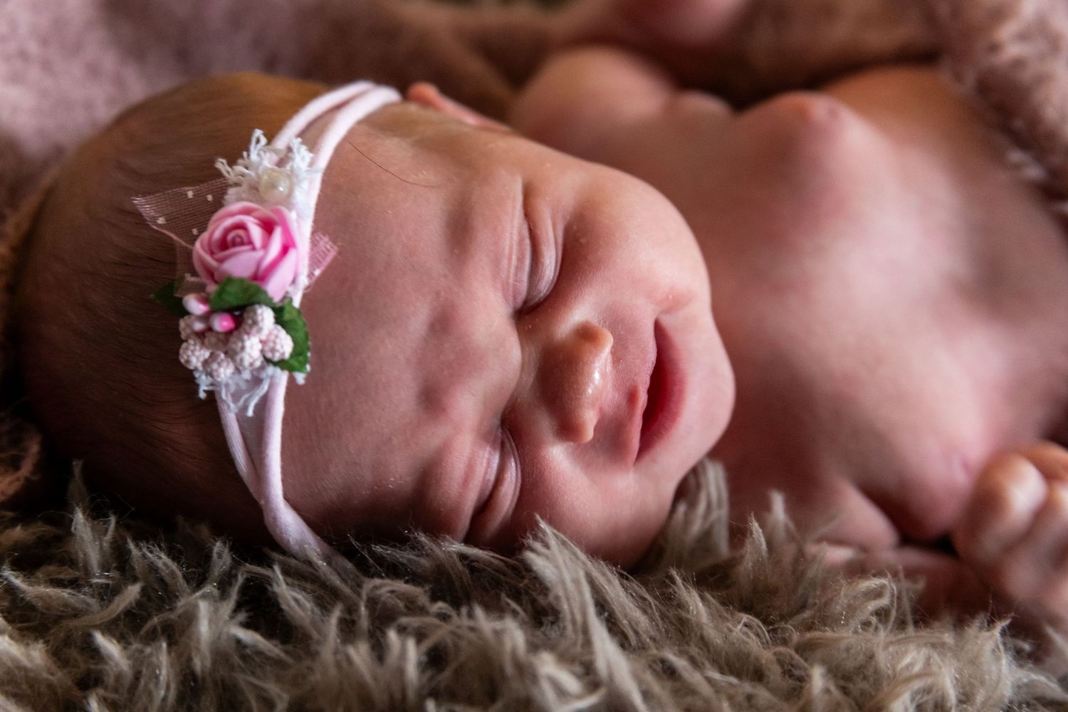 Sleeping newborn wearing delicate pink floral headband rests on soft furry blanket.
