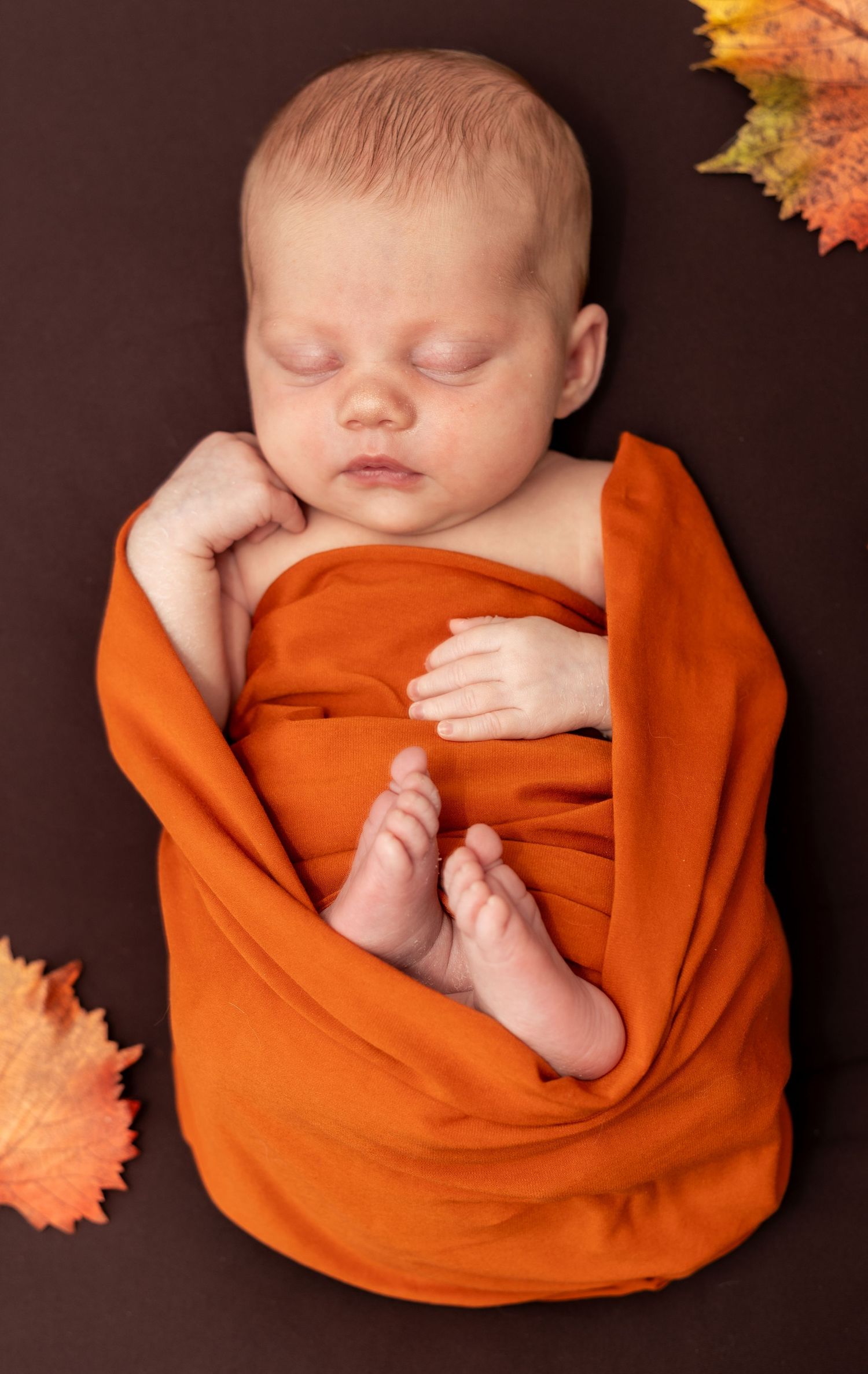 Newborn baby sleeping peacefully wrapped in bright orange swaddle with autumn leaves.