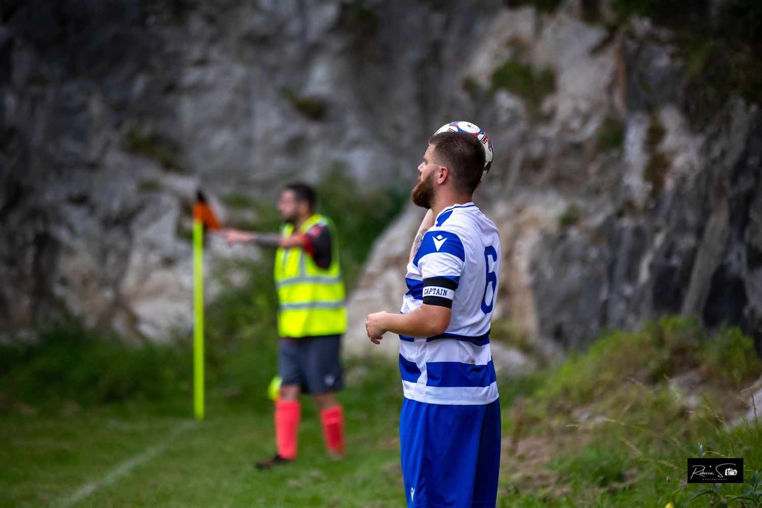 Soccer player in striped jersey standing near rocky cliff with linesman in background.