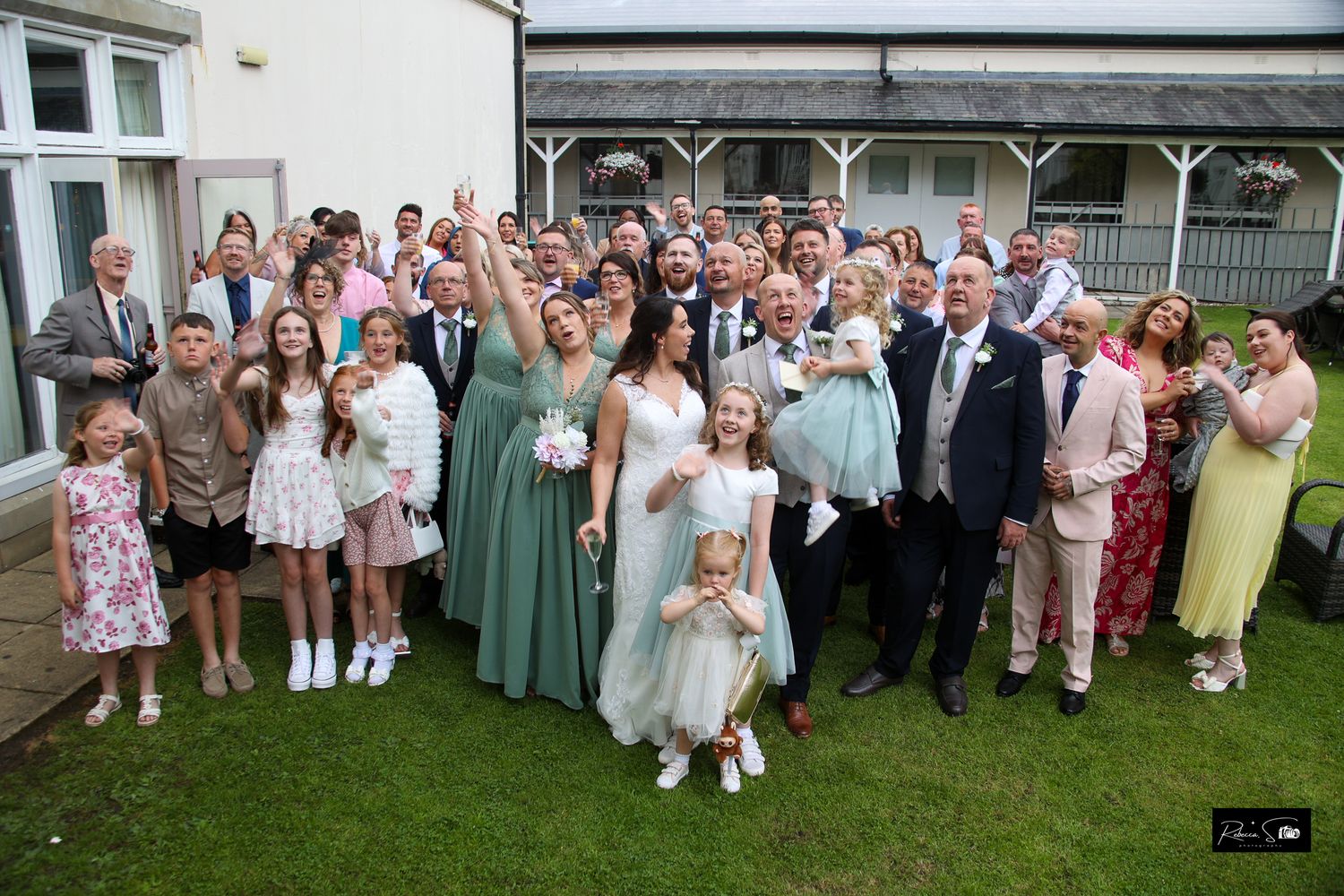 Large group wedding photo with guests gathered on lawn outside venue.