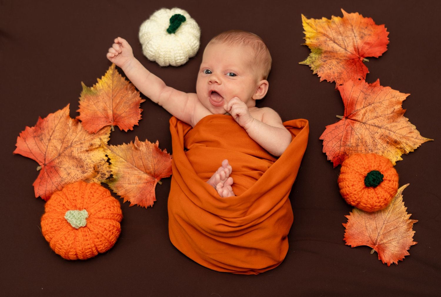 Baby in orange outfit with autumn decorations and pumpkins on dark background.