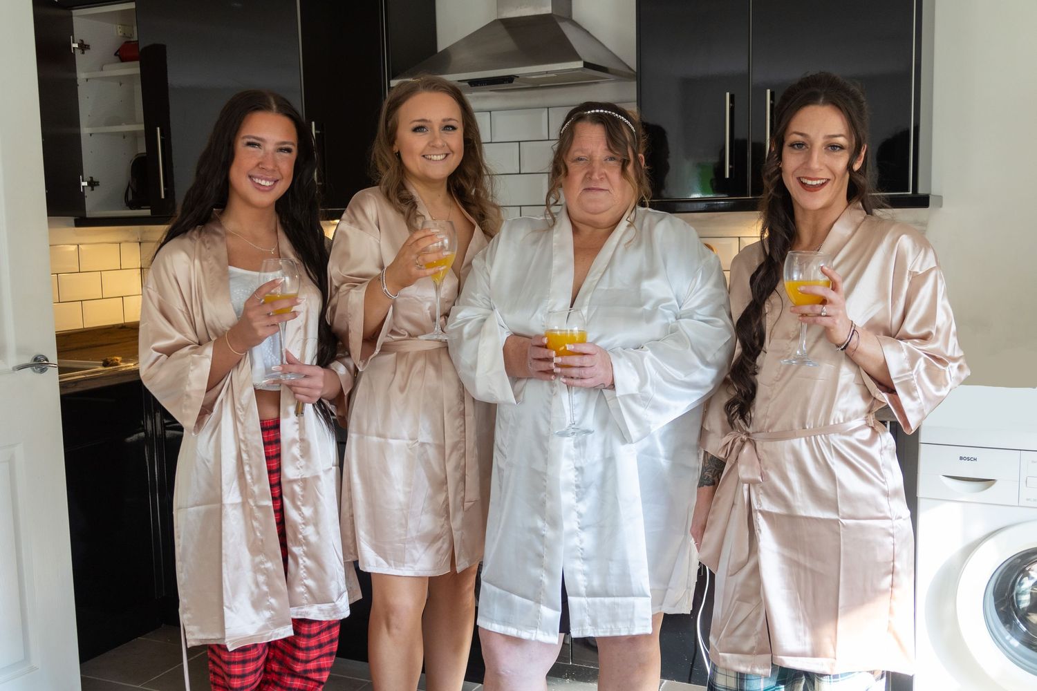 Group of women in matching pink satin robes enjoying drinks in a kitchen setting.