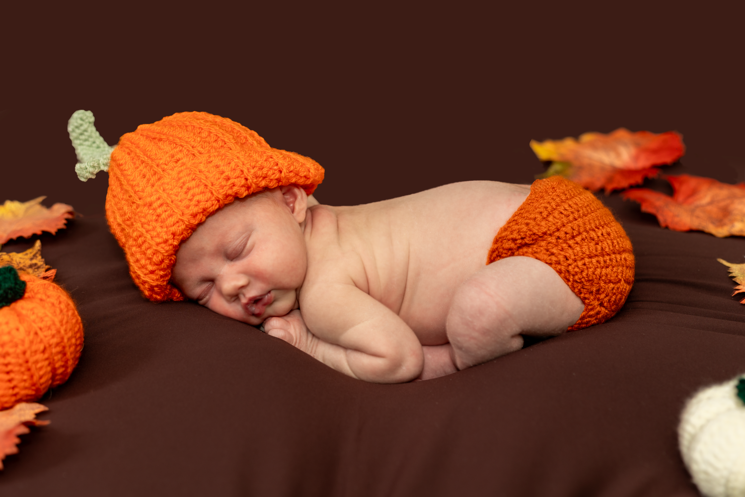 Newborn baby wearing orange knit outfit lying among autumn decorations.