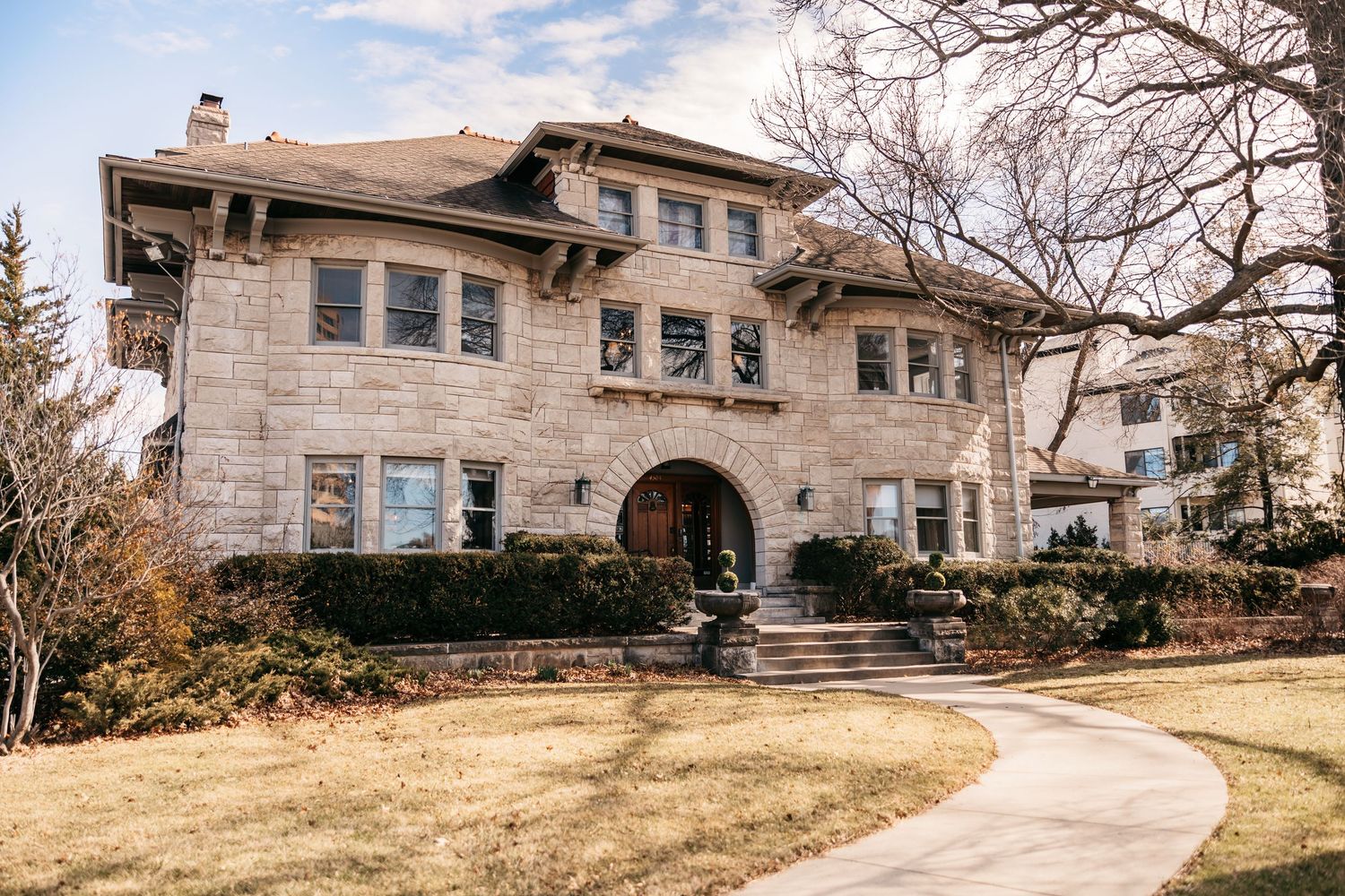 Abandoned stone ranch house with boarded windows sits on a brown winter lawn under cloudy skies.