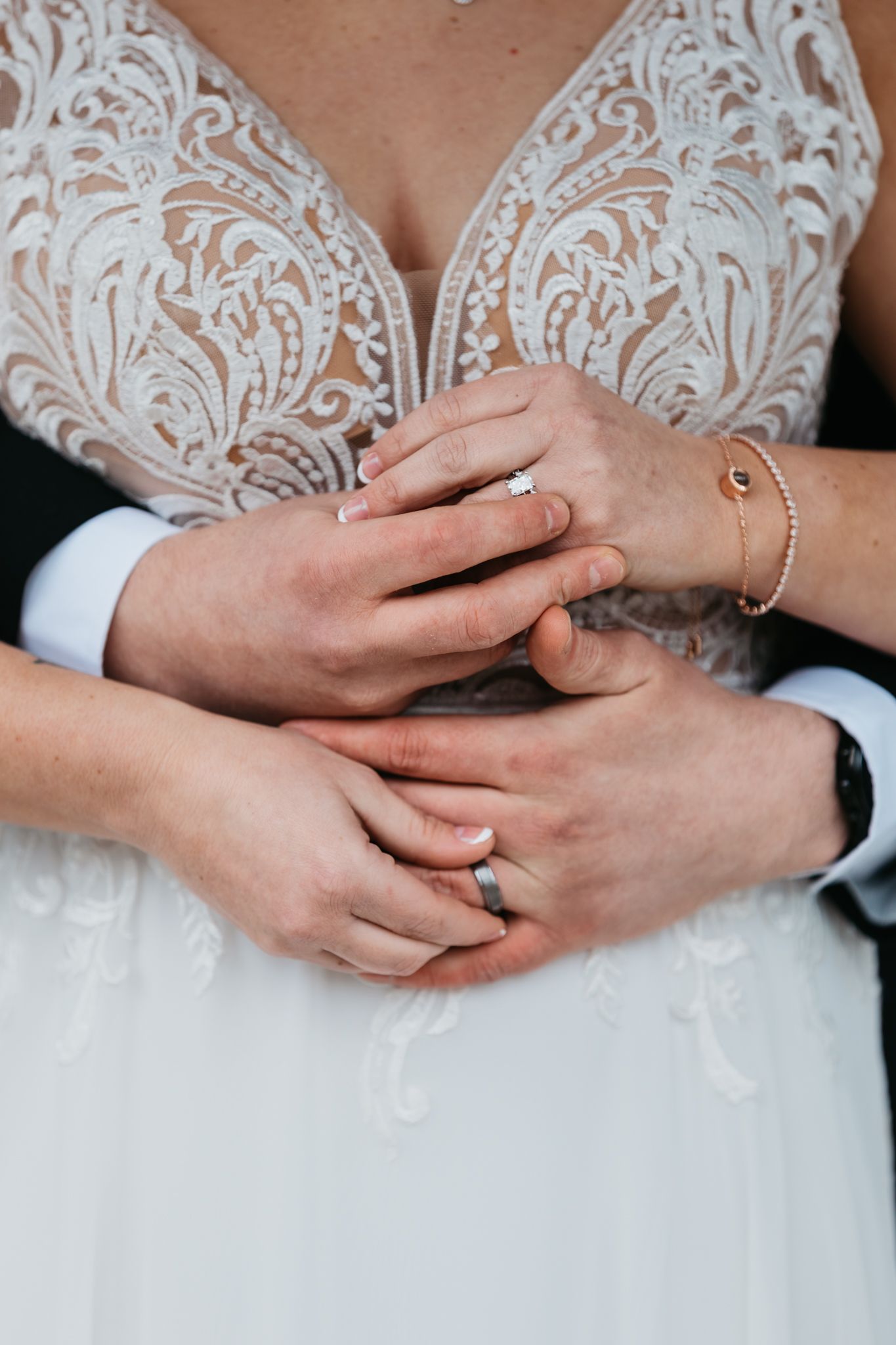 Close-up of hands with wedding rings intertwined against white lace dress.