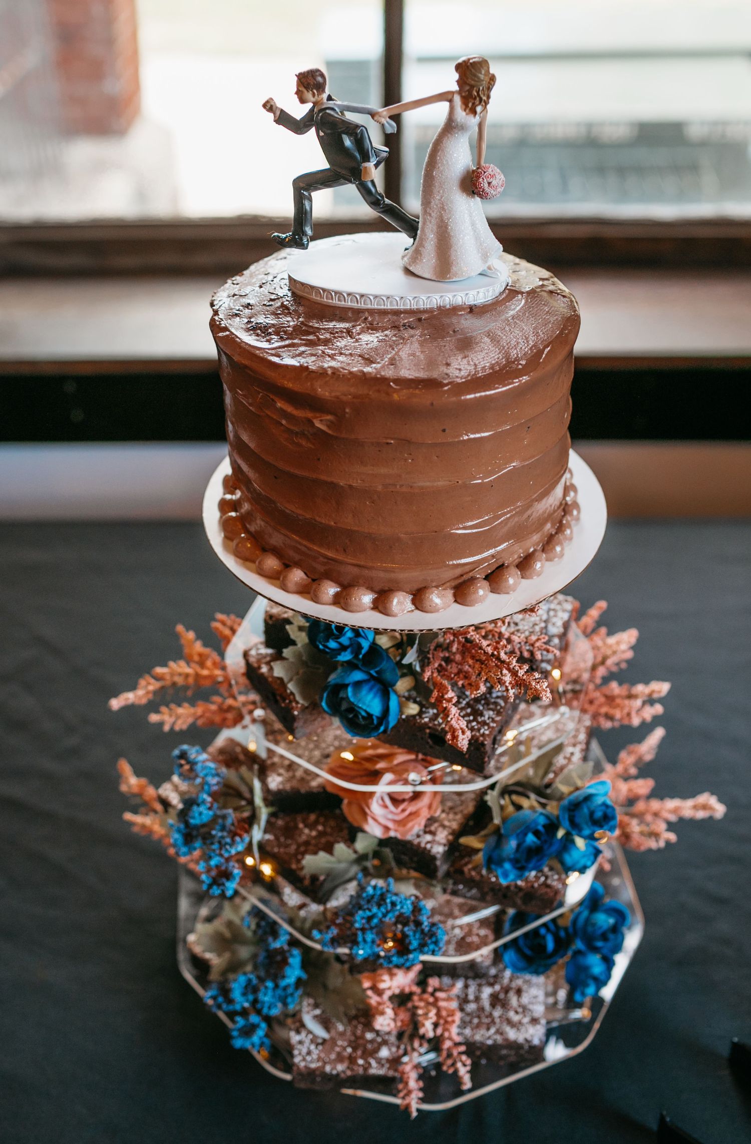 Chocolate layer cake decorated with blue and coral flowers on a rustic cake stand.