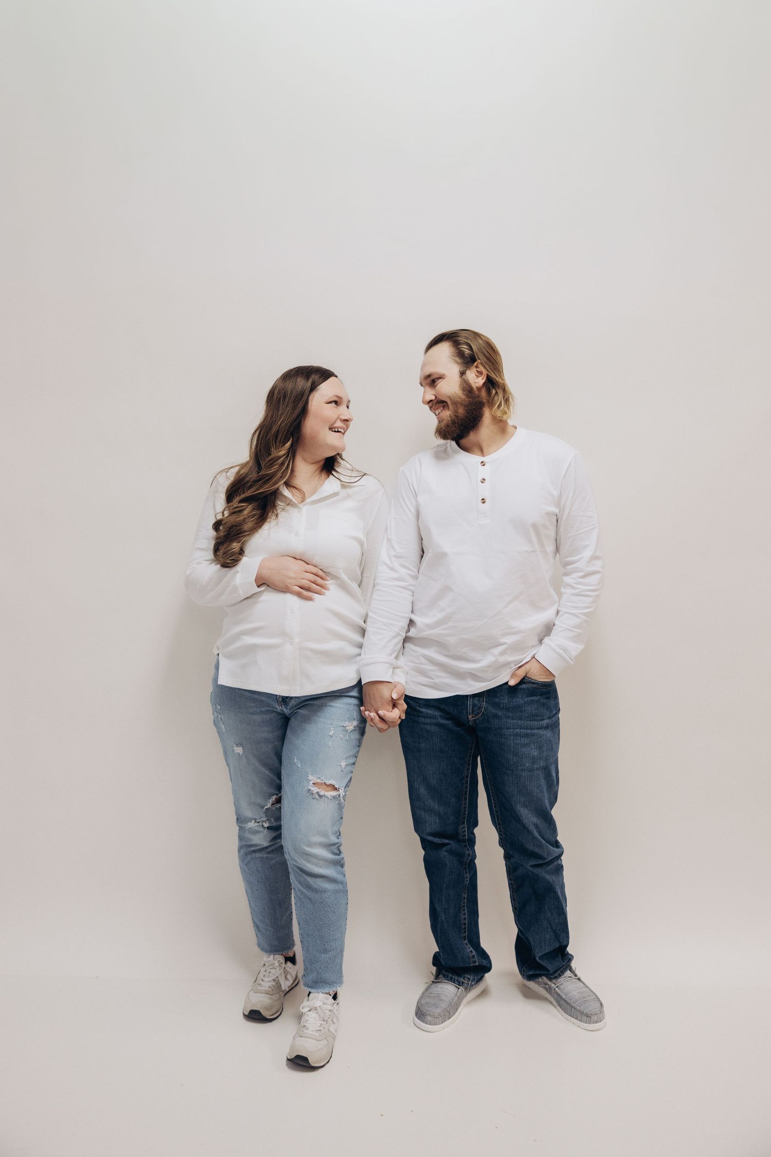 Expecting couple in matching white sweaters sharing loving moment against white backdrop.