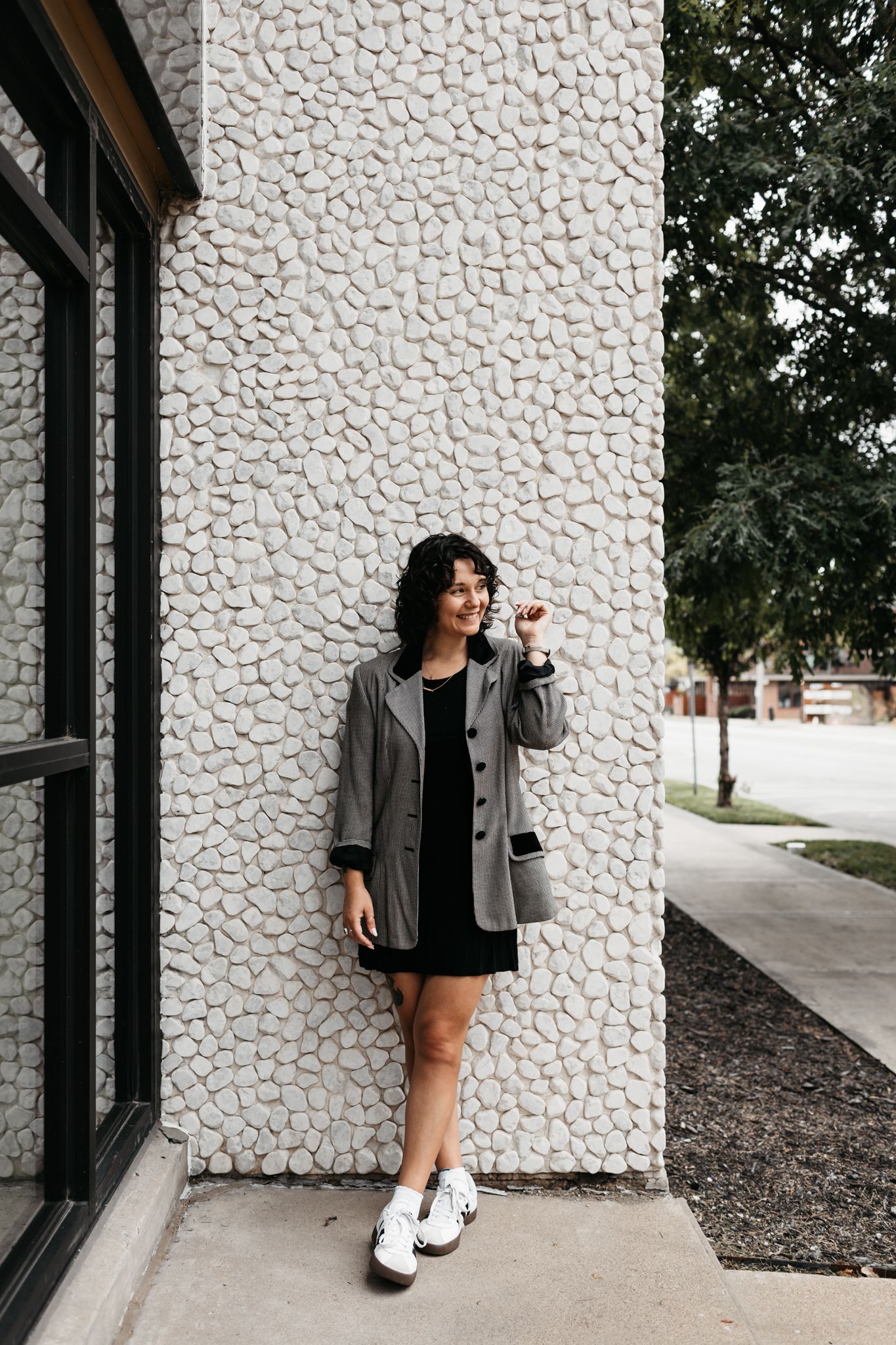 A casual street style look featuring a gray blazer, black dress, and white sneakers against a textured wall.