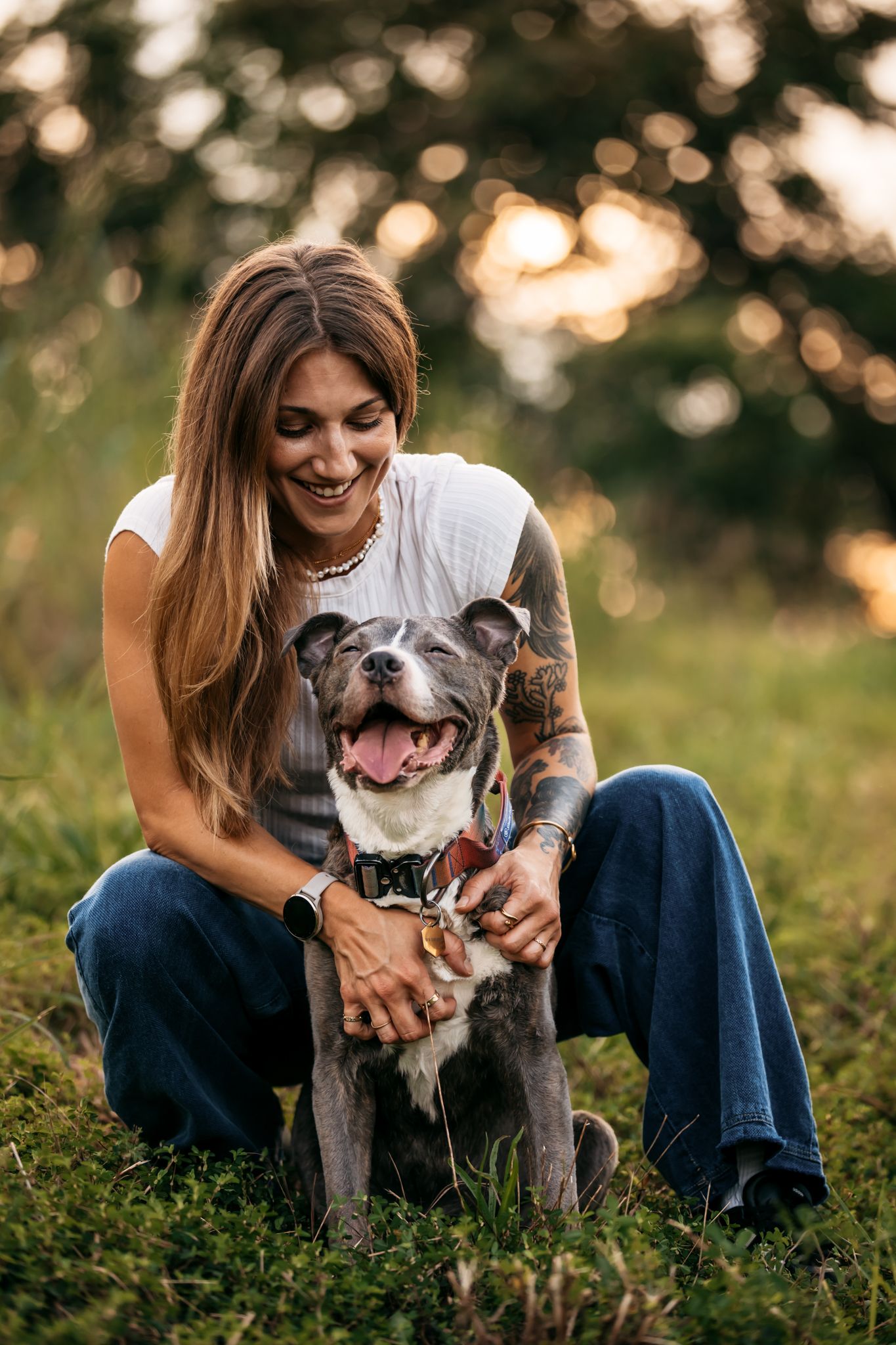 Smiling person cuddling with happy grey pit bull dog in grassy field at sunset.