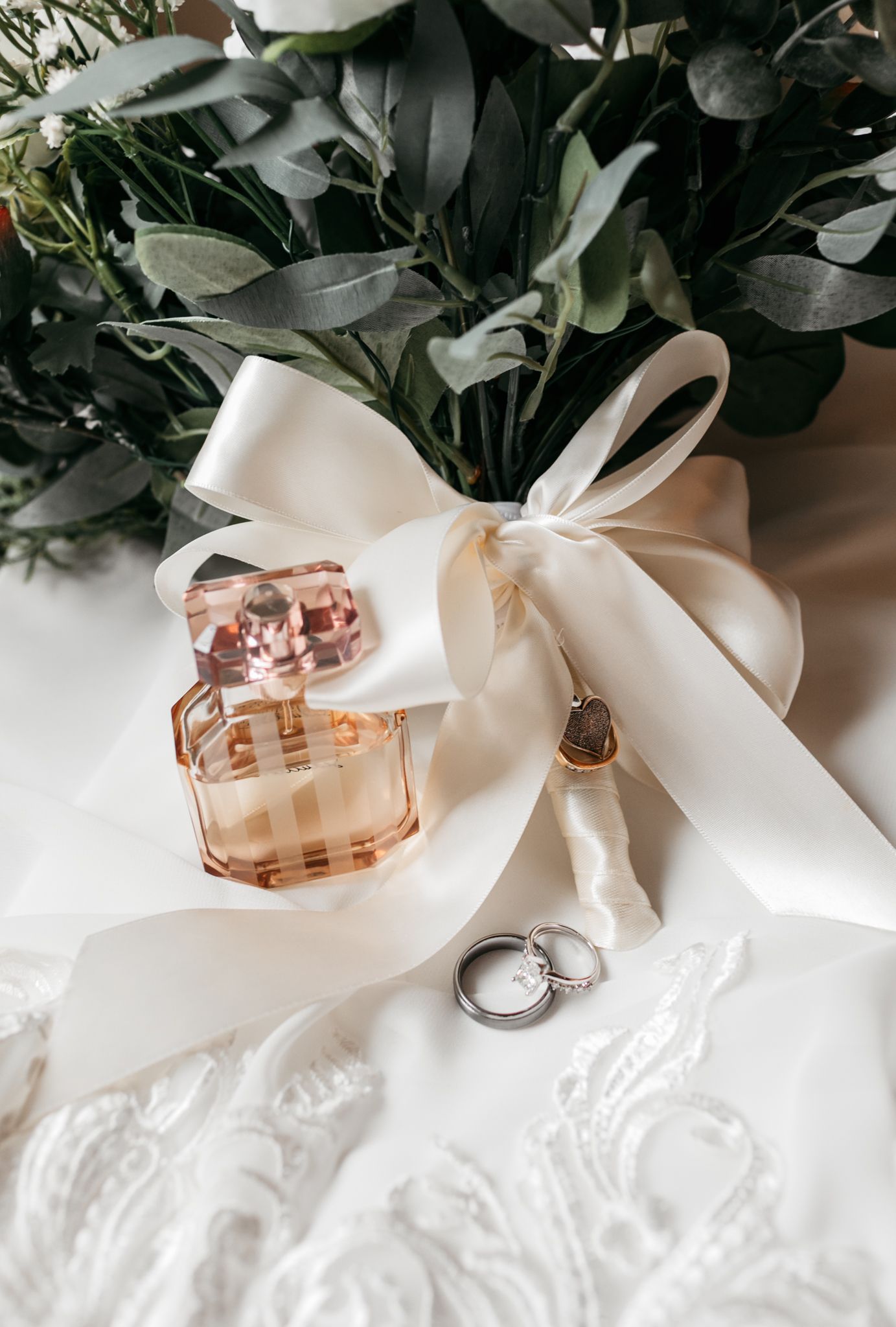 Wedding rings and perfume bottle arranged with white ribbon and greenery.