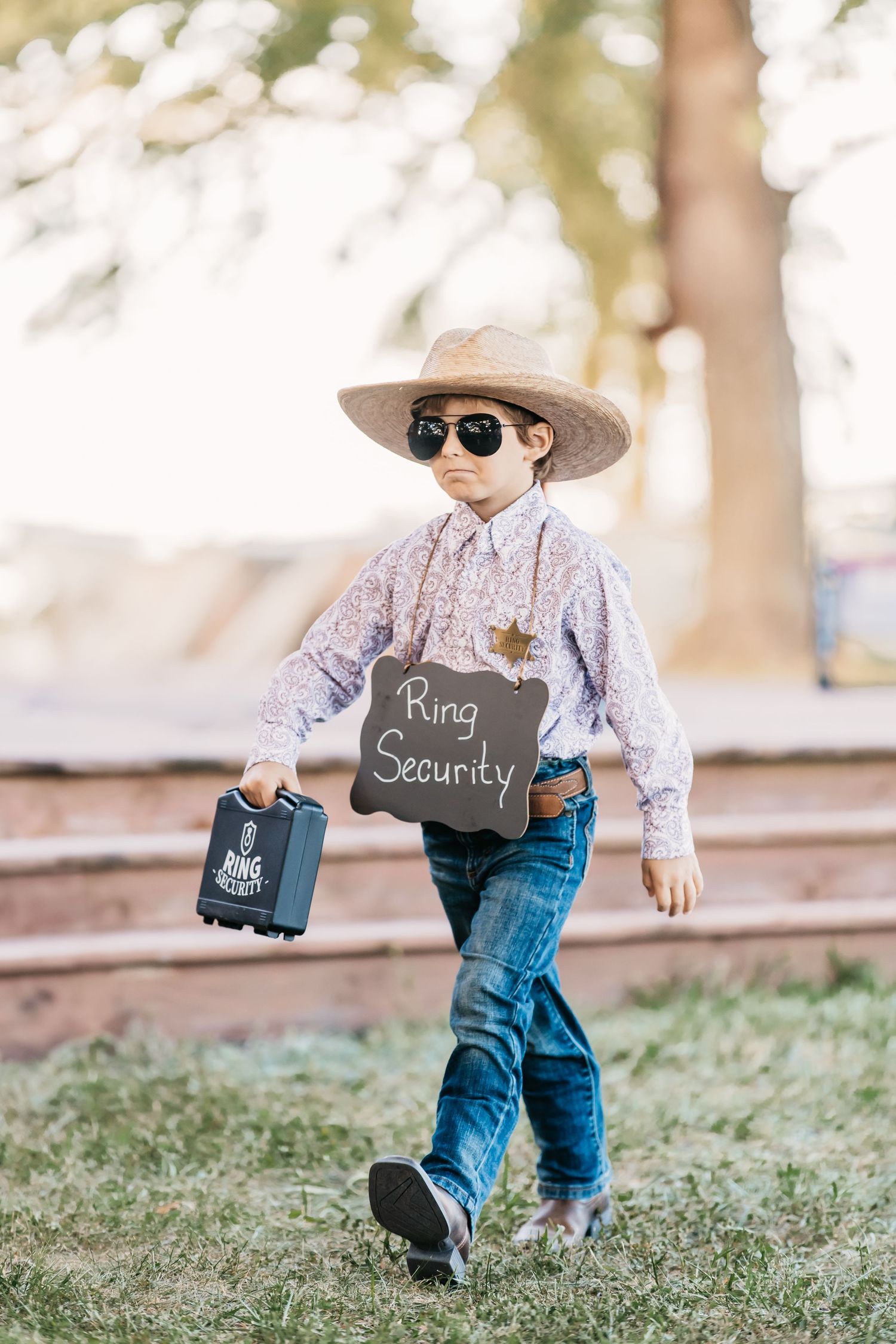 Young child wearing cowboy attire and sunglasses walking through a grassy outdoor setting.