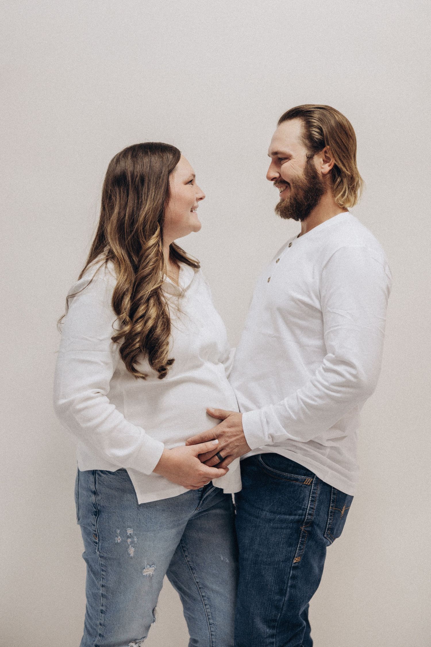 Couple in matching white tops and jeans share intimate moments in minimalist photo series against white wall.