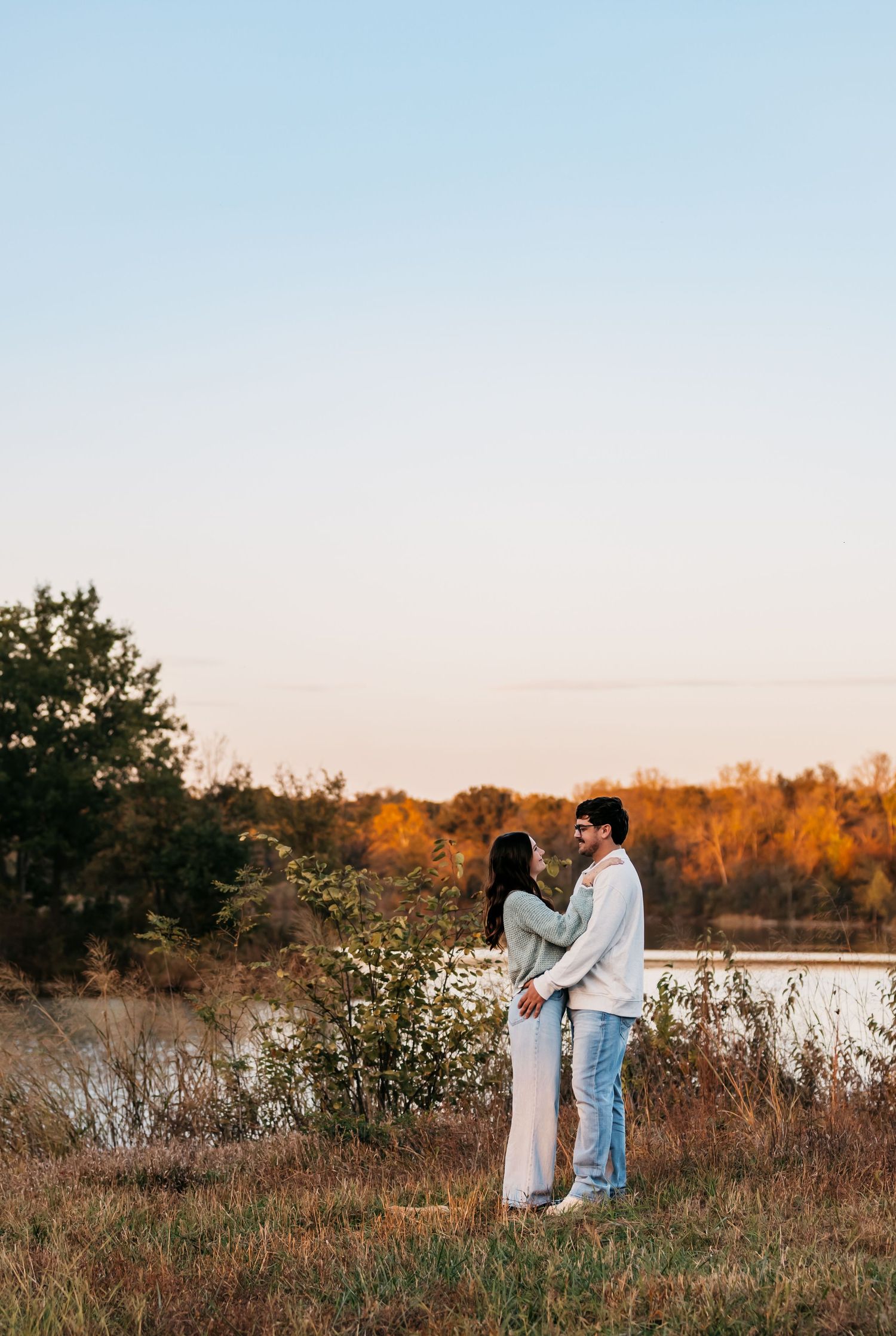 Couple standing together by a lake during autumn sunset with fall foliage in the background.