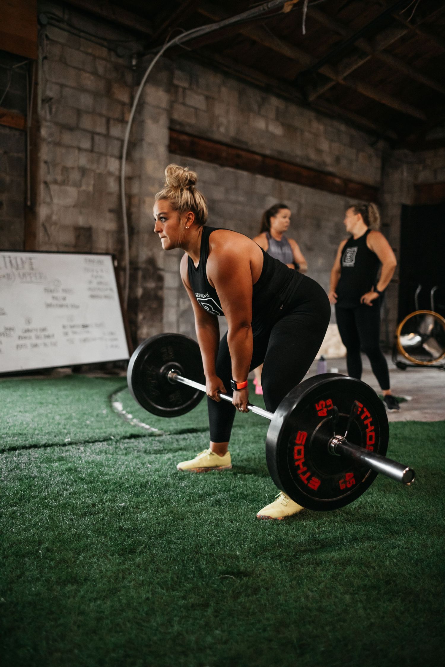 Athletes performing barbell exercises on artificial turf in an industrial gym setting.