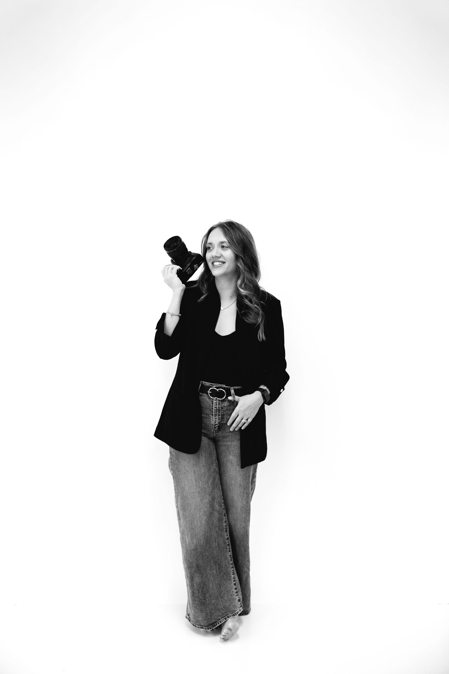 Black and white portrait of photographer holding camera wearing blazer and jeans.