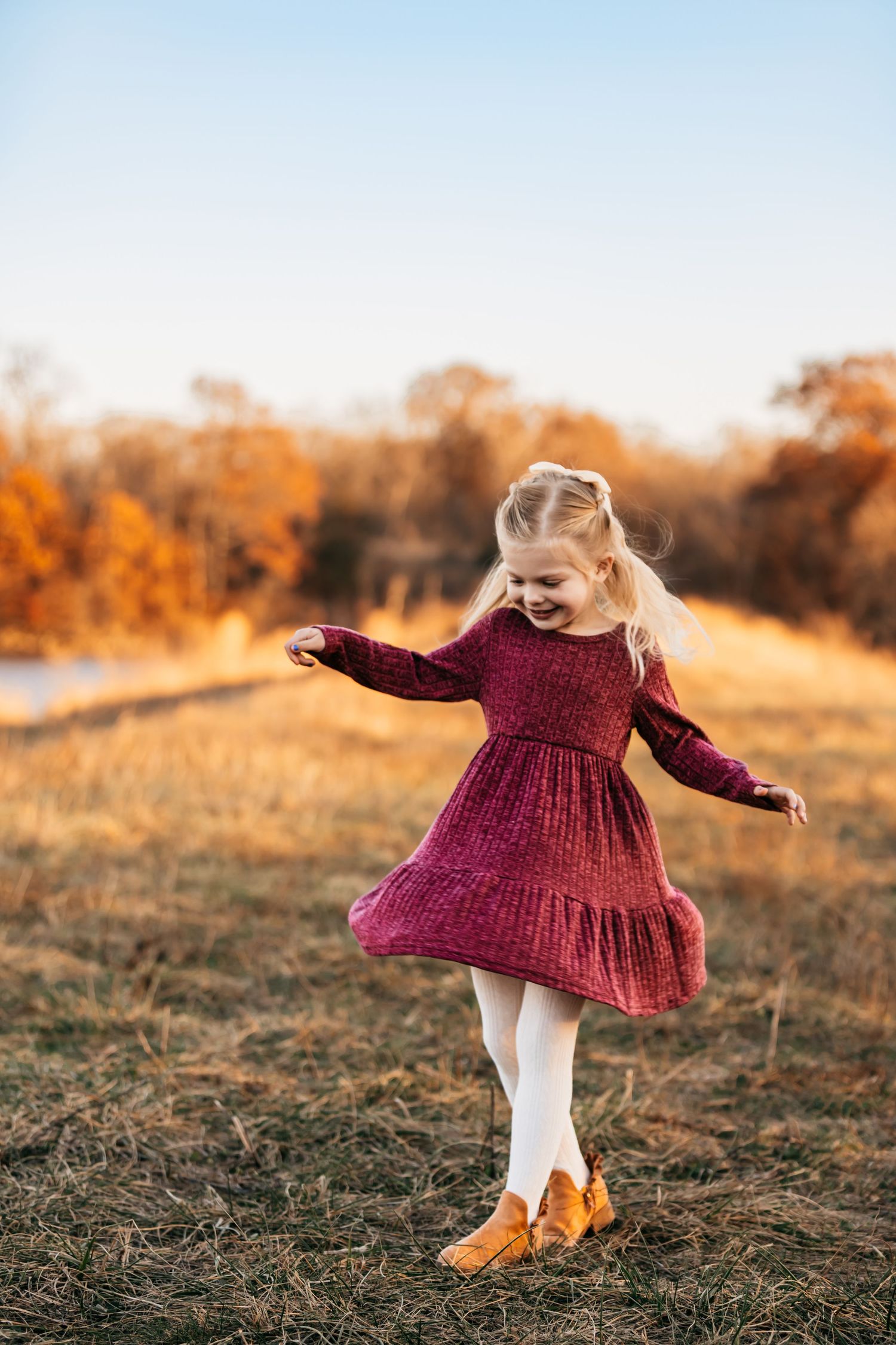 Child in burgundy dress twirling in a golden autumn field at sunset.