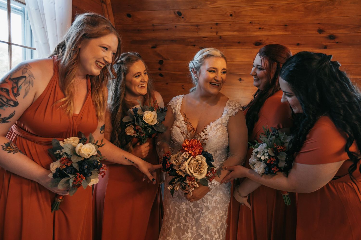 Bridesmaids in burnt orange dresses celebrate with bouquets in wooden interior.