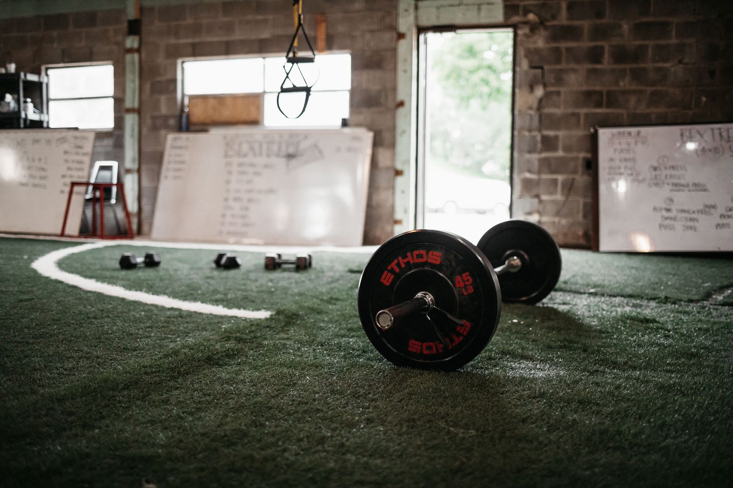 Barbell and weight plates arranged on artificial turf in an industrial-style gym setting.