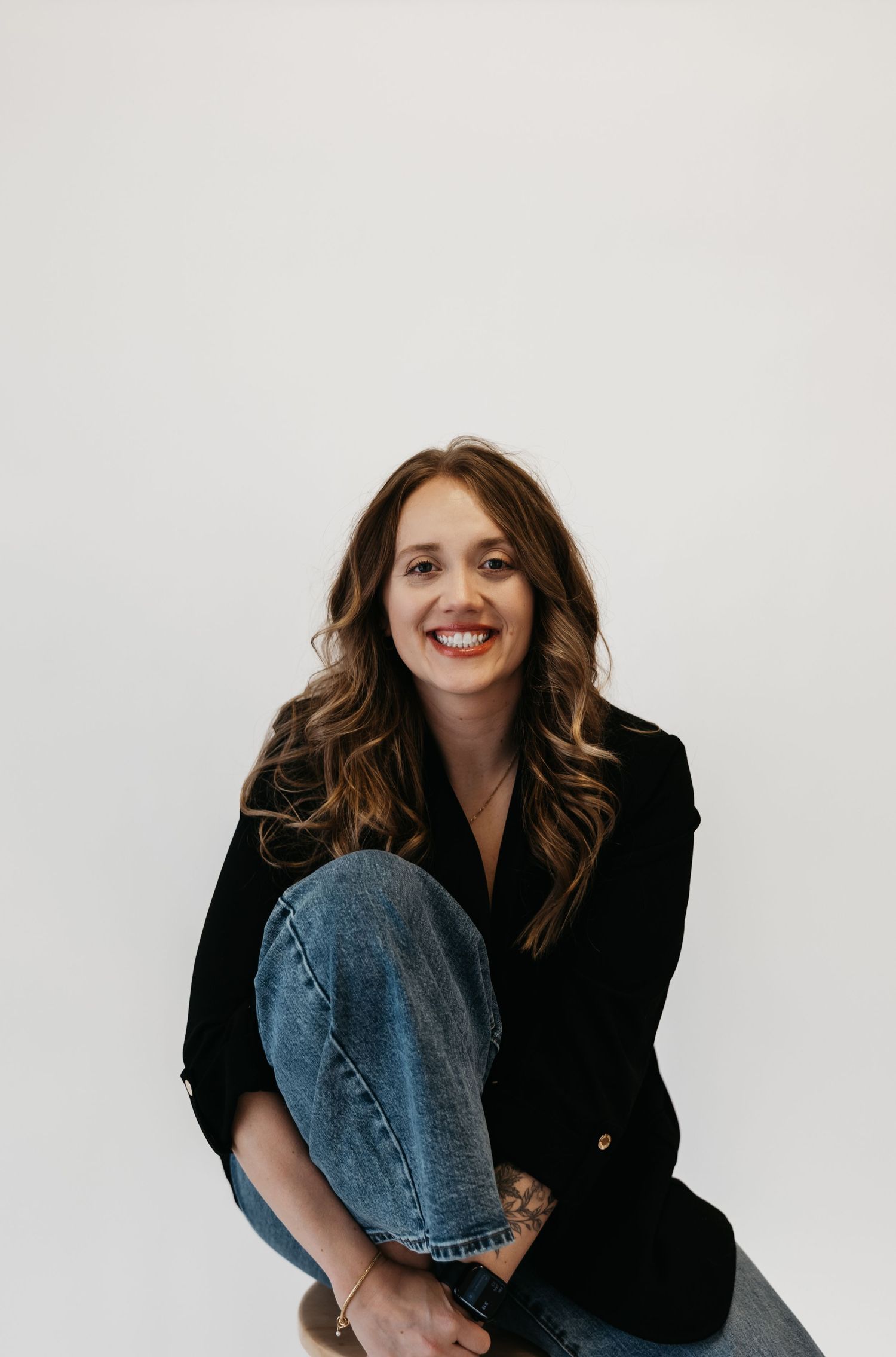 Professional portrait of someone in a black blazer and jeans against a white background showing a bright smile.