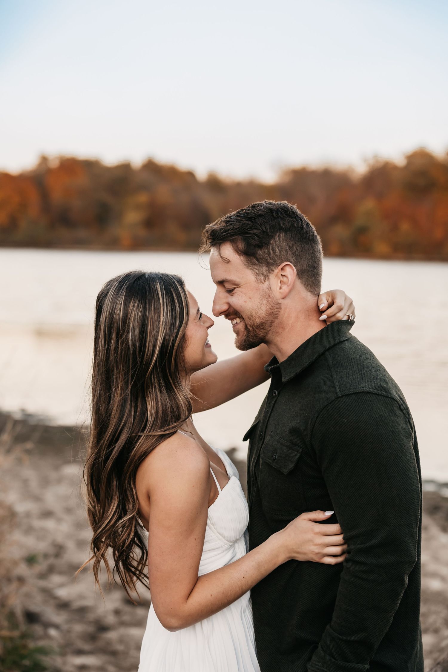 Romantic couple embracing by a lake during sunset with fall foliage in background.
