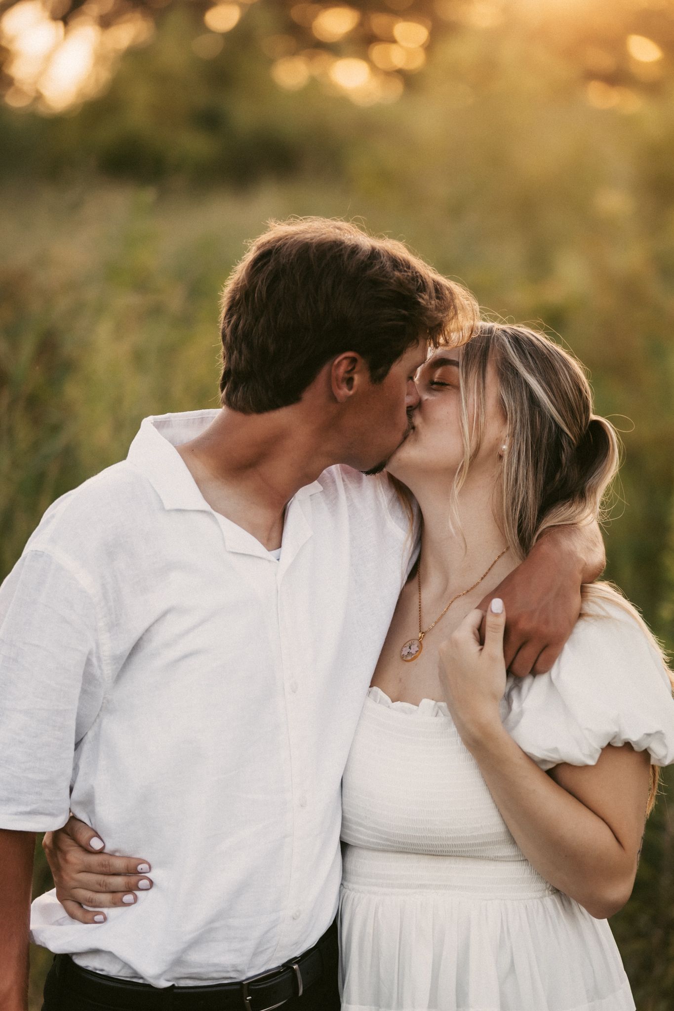 Romantic couple sharing intimate kisses in a golden sunset field wearing white clothing.