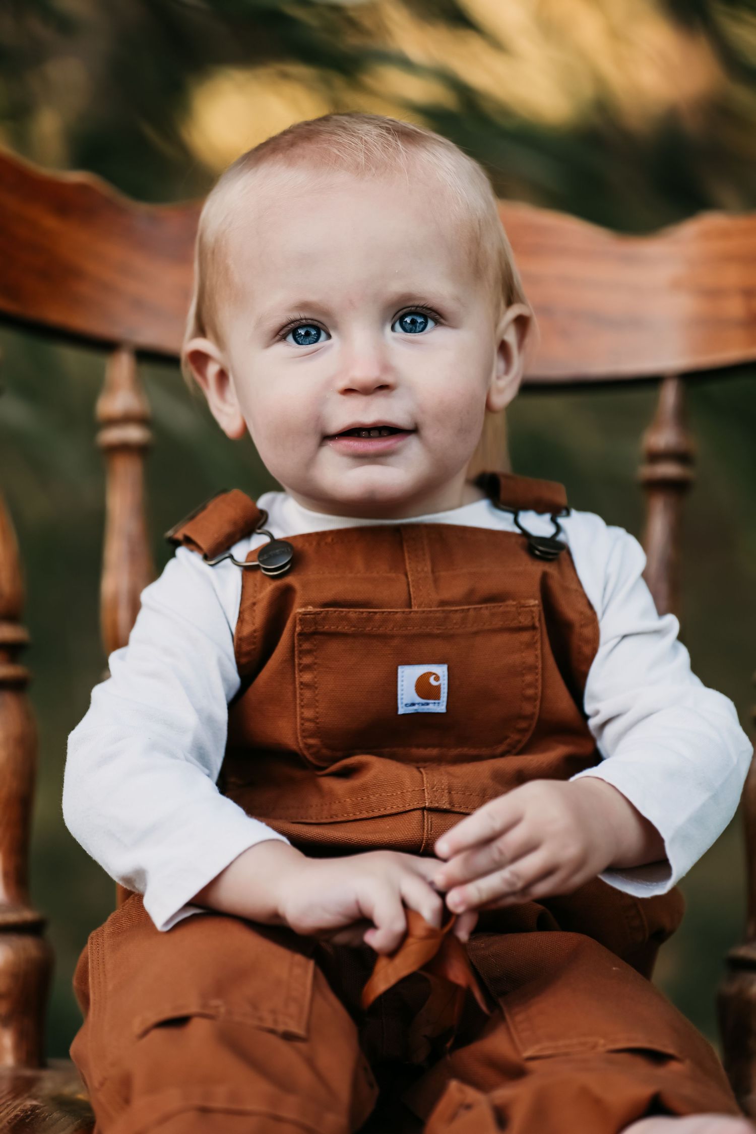Baby wearing brown overalls and white shirt smiles while sitting in wooden chair.