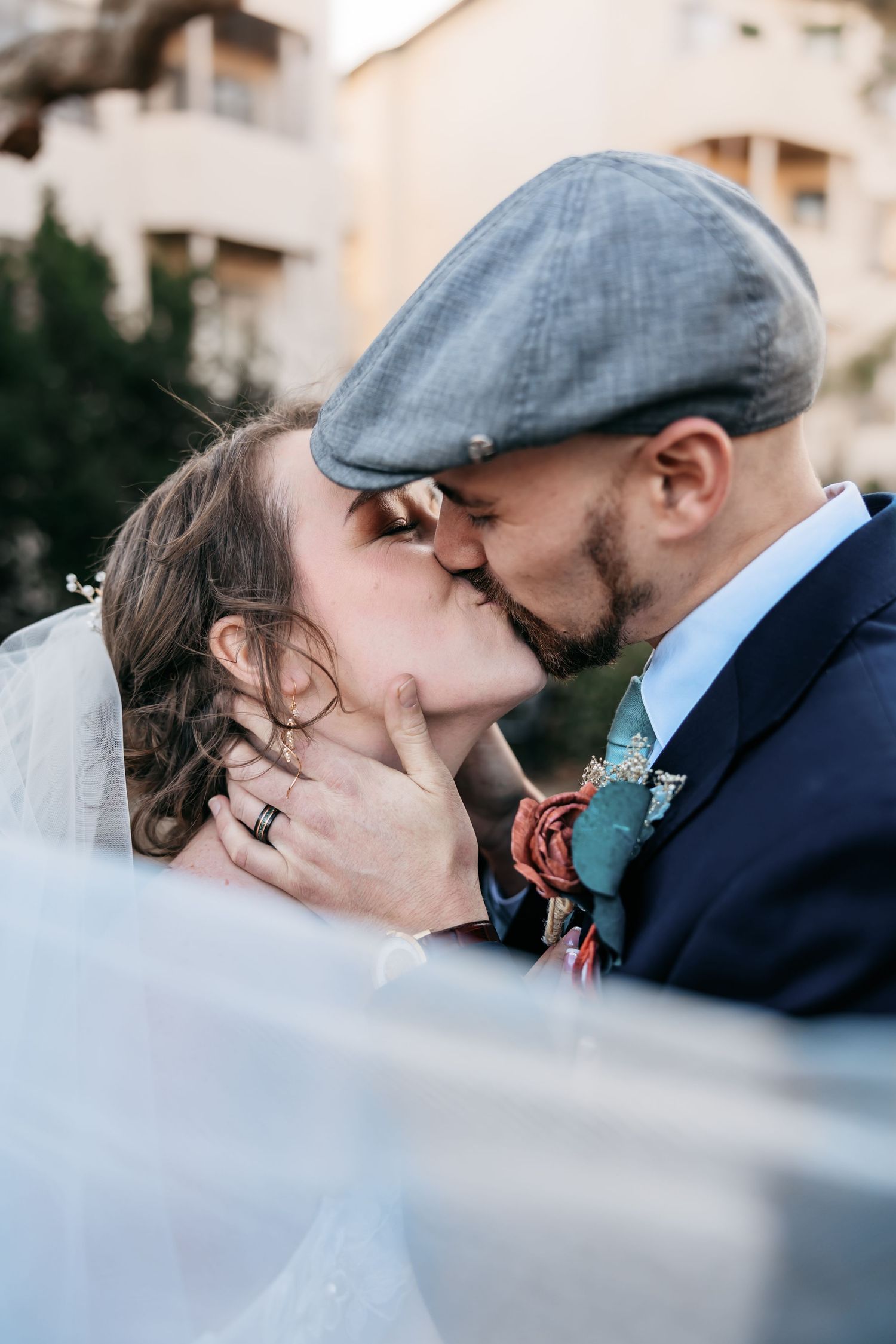 A romantic wedding kiss captured in an intimate moment with the groom wearing a flat cap and navy suit with teal boutonniere.