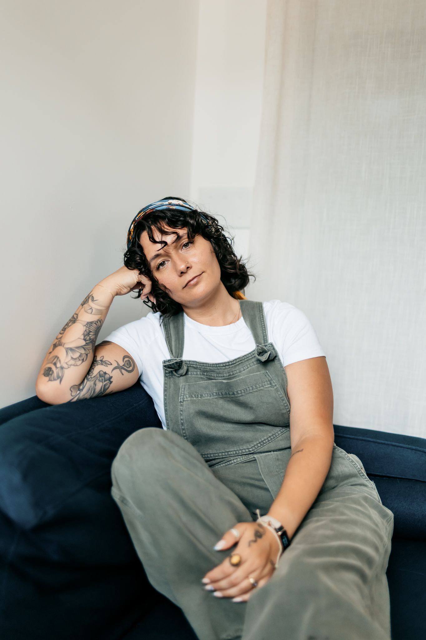Casual portrait of someone relaxing on a couch wearing olive overalls and a white t-shirt with visible tattoos.