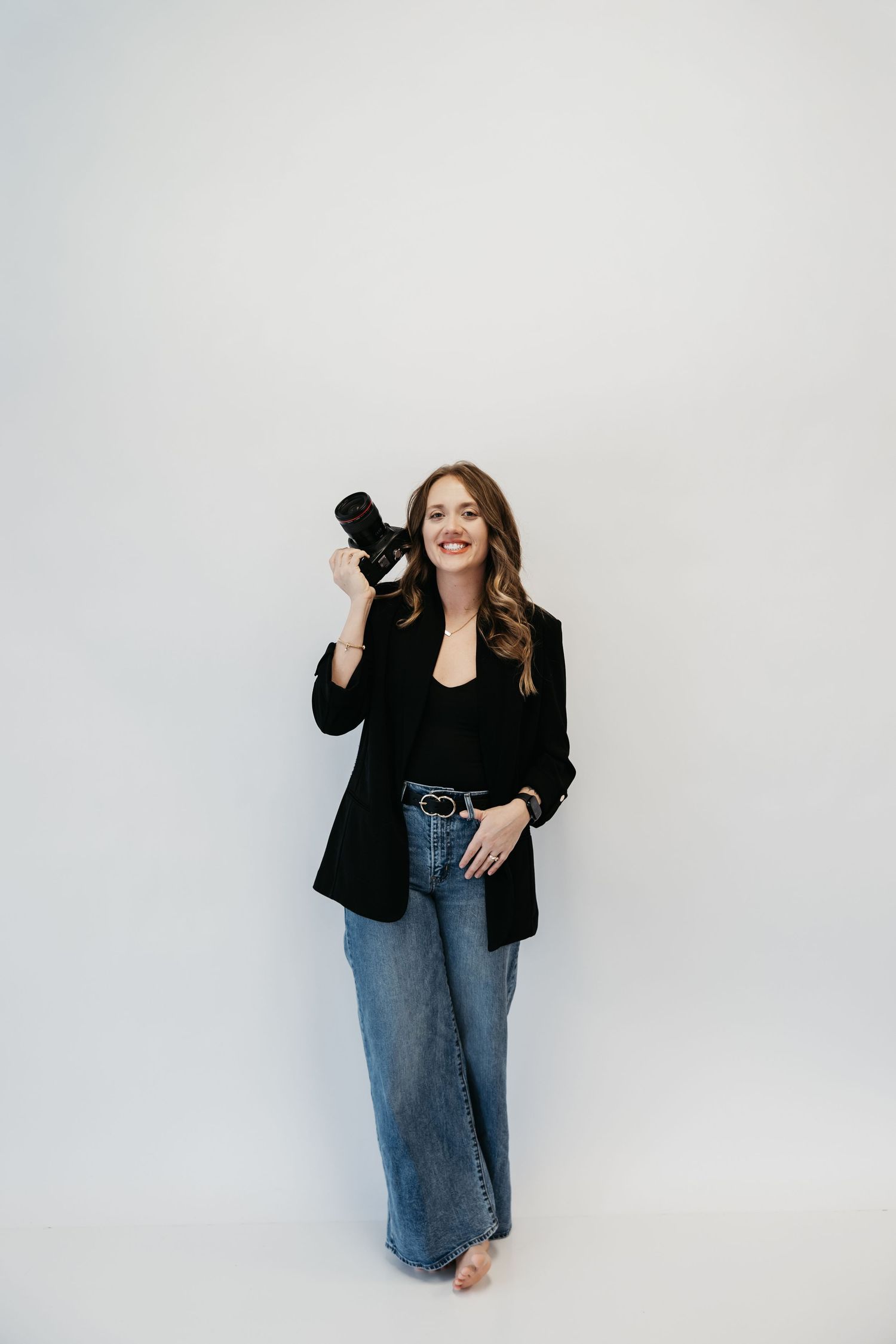 Studio portrait against white background holding camera wearing black blazer and wide-leg jeans.