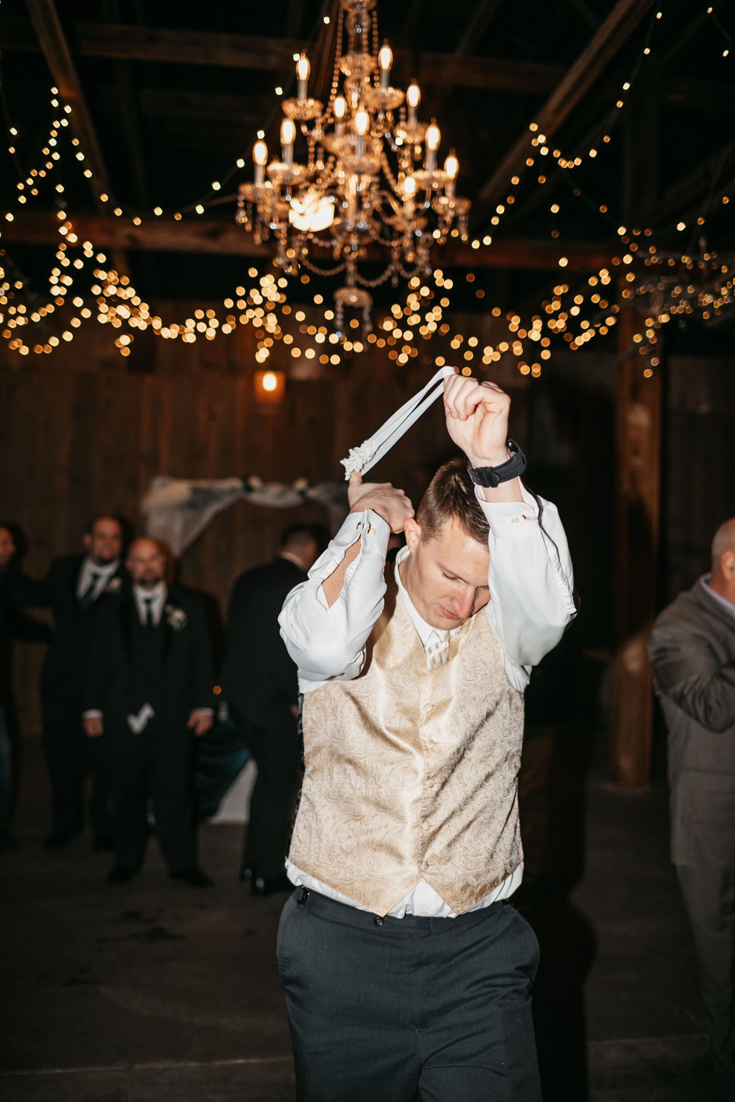 Wedding guests dance energetically under twinkling chandeliers at a rustic barn reception.