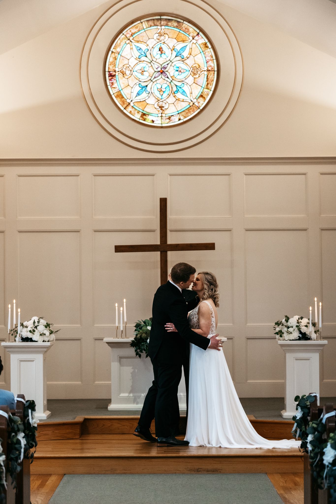 Wedding ceremony in a chapel with stained glass windows and white paneled walls.