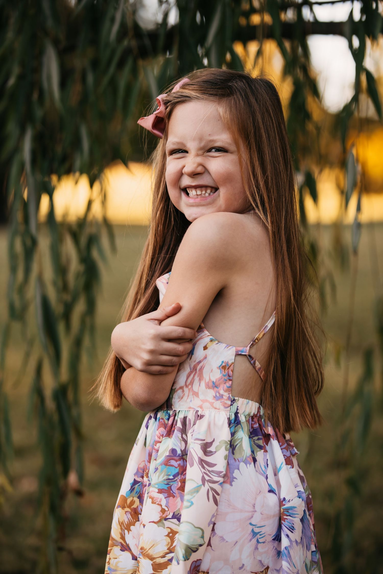 Young girl in floral dress laughing joyfully among willow branches at sunset.