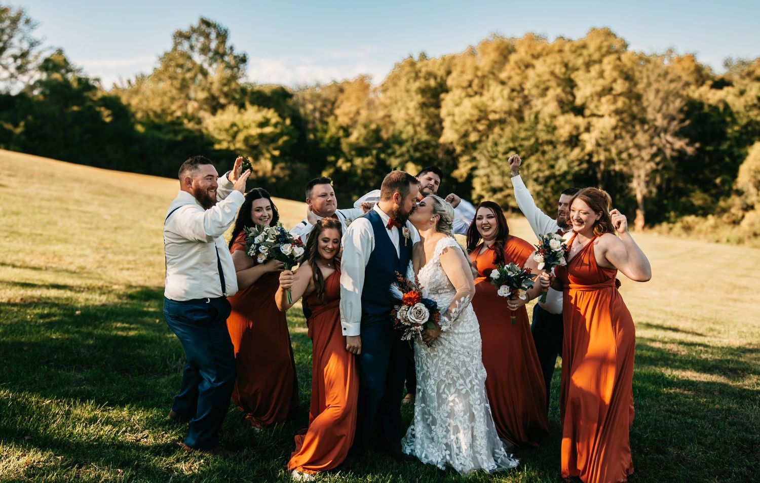 Wedding party in orange and floral dresses pose together in a sunny outdoor field.