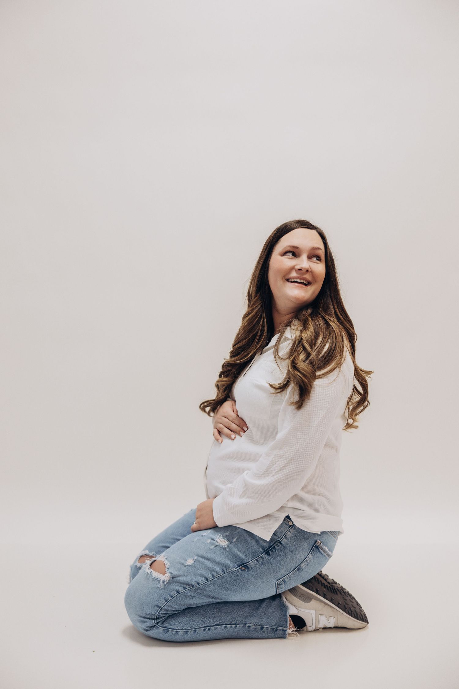 Expectant mother in casual wear kneeling and smiling against white background.