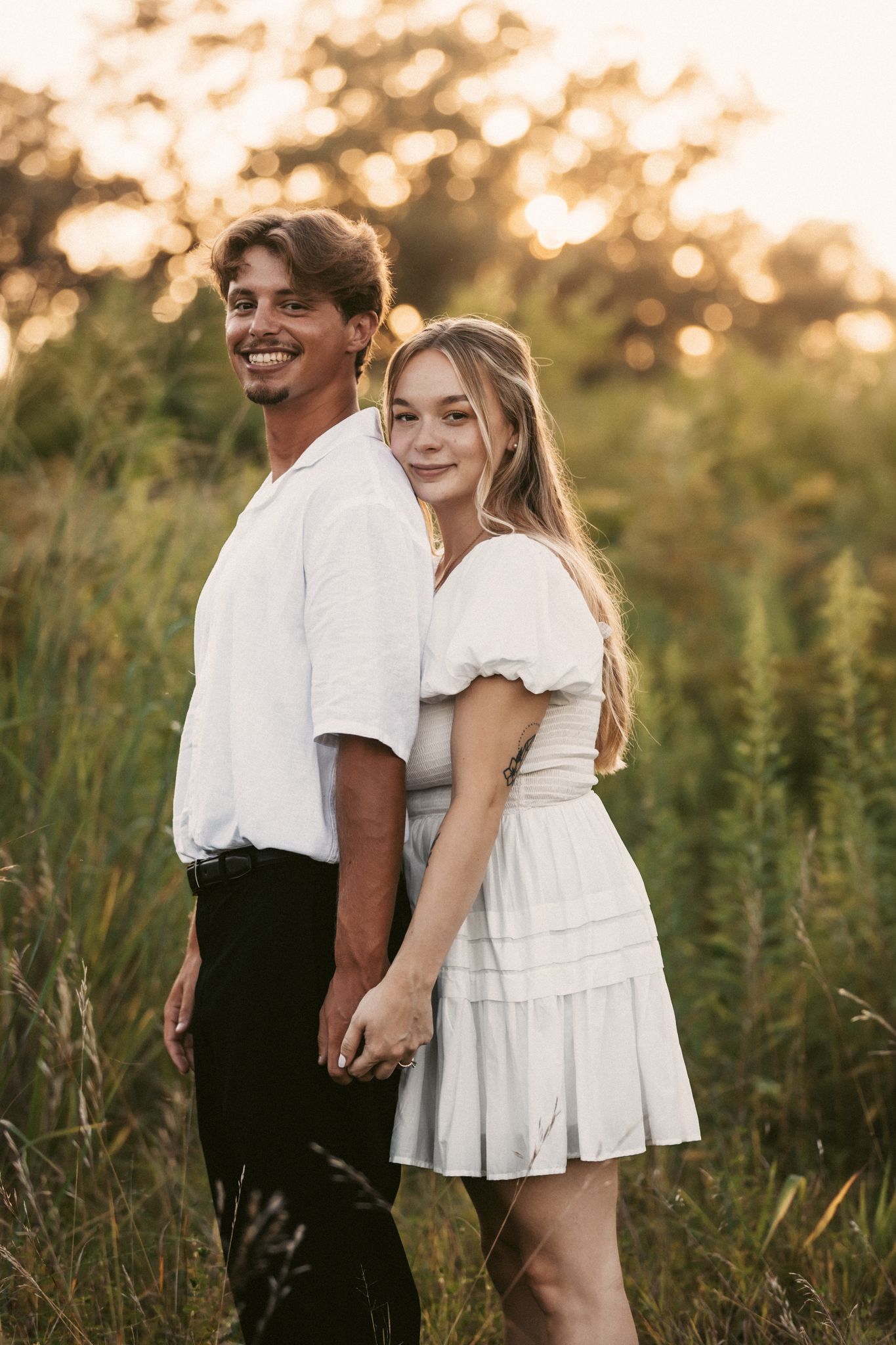 Couple in coordinated white and black outfits posing in tall grass during sunset.